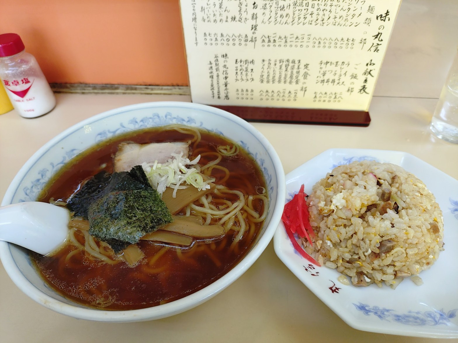 丸信の醤油ラーメンと半チャーハンが並ぶテーブル。奥に手書きの品書き、塩入れとグラスが見える。